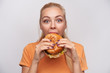 Close-up of overjoyed young blue-eyed cute blonde female eating tasty burger with great pleasure and looking happily at camera with wide eyes opened, isolated over white background