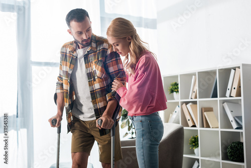 Fotografía handsome man holding crutches while standing near woman at home