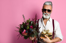 Stylish Middle-aged Bearded Man With A Modern Haircut, Sunglasses And Fashionably Dressed Holds A Bouquet Of Flowers And A Gift. Going On A Date