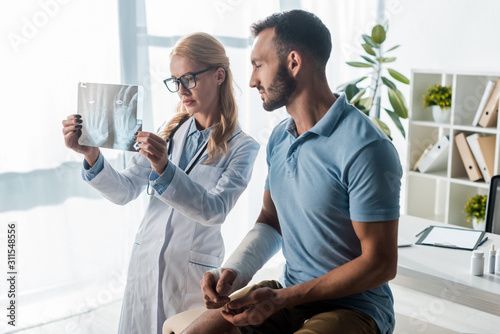 Fotografía attractive orthopedist in glasses holding x-ray near handsome injured man