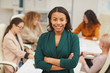 Charming African American woman standing with arms closed looking at camera smiling with colleagues coworking behind her, waist up portrait