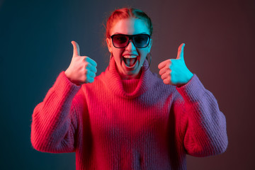 Wall Mural - Showing thumbs up. Caucasian woman's portrait isolated on gradient studio background in neon light. Beautiful female model with sunglasses, red hair. Concept of human emotions, facial expression, ad.