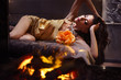 A girl in sexy silk pajamas lies on a bed against a blazing fire in a fireplace.
