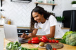 Smiled pretty girl is looking on the laptop screen  on the modern kitchen on the table full of vegetables and fruits, dressed in white t-shirt