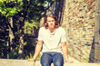 © Alexander Image - Portrait of Young Handsome American Man with long dark brown hair in New York City, wearing white short sleeve shirt, blue jeans, sitting on rocks half wall under sun in summer, thinking, relaxing..