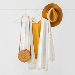 Wall Mural - White knitted jumper on hanger with brown hat and bamboo bag on white background. Elegant fashion outfit. Spring wardrobe. Minimal concept.