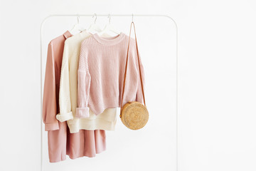 Wall Mural - Feminine warm sweaters and pale pink dress with bamboo bag on hanger on white background. Elegant fashion outfit. Spring wardrobe. Minimal concept.