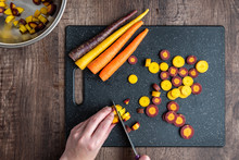 Woman’s Hands Cutting Rainbow Carrots On A Black Cutting Board, Stainless-steel Bowl, Wood Table