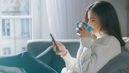 Beautiful Young Woman Using Smartphone, Drinks Tea while Sitting on the Chair. Sensual Girl Wearing Sweater Surfs Internet, Posts On Social Media, Shares Pictures while Relaxing in Cozy Apartment