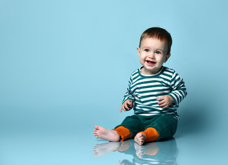little baby boy in stylish casual clothing barefoot sitting on floor and smiling over blue wall back