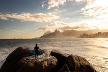 Surfer With Surfboard In Hand Watching Sunset At Pedra Do Arpoador, Arpoador Beach, Rio De Janeiro, Brazil