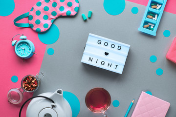 Healthy night sleep creative concept. Sleeping mask, alarm clock, earphones, earplugs and pills. Split two tone, pink and grey paper background with circles and tea. Lightboard with text 