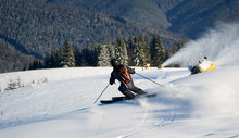 Back View Of Man Skiing On Prepared Hill With Fresh New Powder Snow. Artificial Snowfall In Sunny Day. Electric Snow Gun Machine. Ski Lesson In Skiing School. Scenic Wooded Mountains On Background.