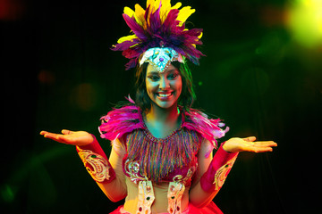 Beautiful young woman in carnival mask and stylish masquerade costume with feathers in colorful lights and glow on black background. Christmas, New Year, celebration. Festive time, dance, party.