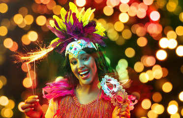 Wall Mural - Beautiful young woman in carnival mask and stylish masquerade costume with feathers and sparklers in colorful bokeh on black background. Christmas, New Year, celebration. Festive time, dance, party.