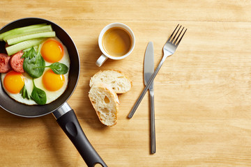 Wall Mural - top view of fried eggs with spinach leaves, cucumber and sausage in frying pan near coffee, bread and coffee on wooden table