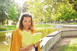 © Mangostar - Pensive woman enjoying reading in park, sitting on parapet near river. Green grass background. Curly haired black woman in casual posing outdoors. Reading book, leisure concept