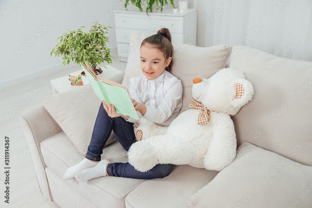Cute little girl sitting on the sofa with teddy-bear near her, reading ...