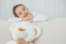 Big White Soft Bear Is On The Sofa. Girl Is Standing Behind The Sofa, Leaning Over Its Back, Folding Her Hands, Looking At The Camera, Smiling Pleasantly.