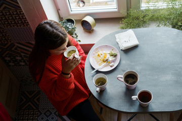 Blonde girl with long hair in a red sweater sitting at the dining table by the window eating breakfast. A few cups of tea and coffee for a family feast. View from above