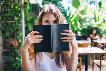 Charming female hiding face behind book in restaurant