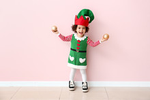 Little Girl In Costume Of Elf And With Christmas Balls Near Color Wall