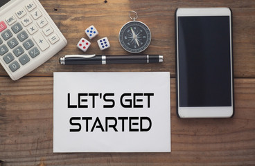 Let's Get Started written on paper,Wooden background desk with calculator,dice,compass,smart phone and pen.Top view conceptual.