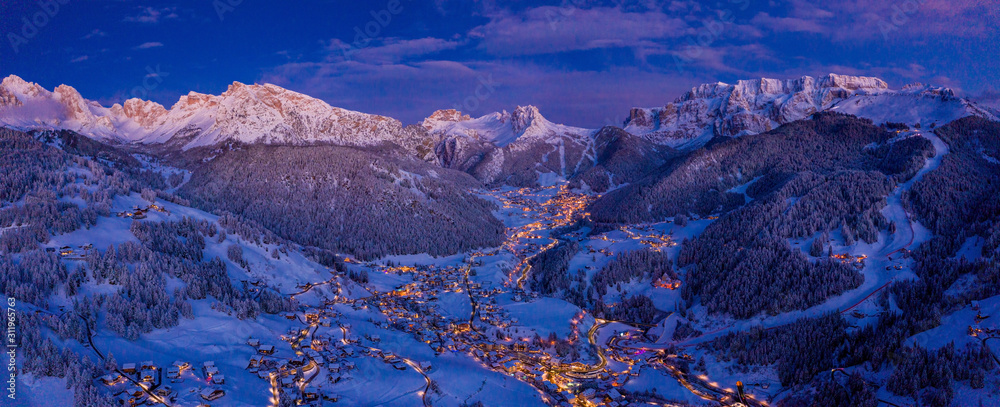 Beautiful panoramic view of Dolomites mountains at dusk during winter time. Magical winter mountain purple sunset with a mountain ski resort village. Christmas time. - obrazy, fototapety, plakaty 