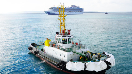 Wall Mural - A bright, powerful, new, industrial tug helps the ship maneuver in port. A tugboat helps moor a cruise ship in the clear blue water of tropical islands on background white cruise ship