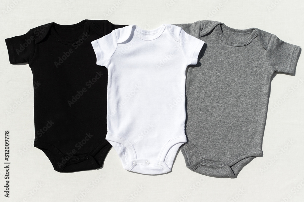 A set of three blank baby bodysuits on a cream textile background in harsh light - Black, white and grey baby grow set mockup -  Styled stock photography - obrazy, fototapety, plakaty 