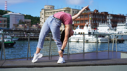 young beautiful flexible girl in the city on the embankment stands in a bridge pose. gymnast in life style clothes at the observation deck in the port does gymnastic elements. backbend pose. yachts