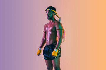 Muay thai. Young man exercising thai boxing on gradient background in neon light. Fighter practicing, training in martial arts in action, motion. Healthy lifestyle, sport, asian culture concept.