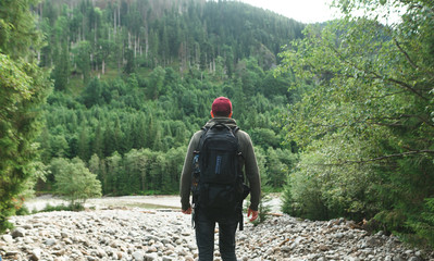 Tourist with backpack standing in mountains near mountain river and looking at beautiful landscape.Man walks in the national park,stands with his back and looks forward. Man and untouched nature.