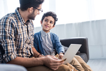 Wall Mural - Jewish father and smiling son using laptop in apartment