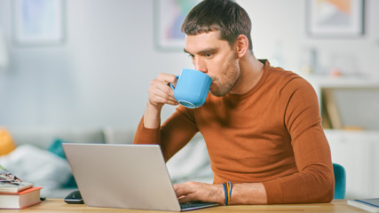 Wall Mural - Portrait of Handsome Smiling Man Working on Laptop, Sitting at His Wooden Desk at Home and Drinking Coffee. Man Browsing Through Internet, Working on Notebook from His Living Room.