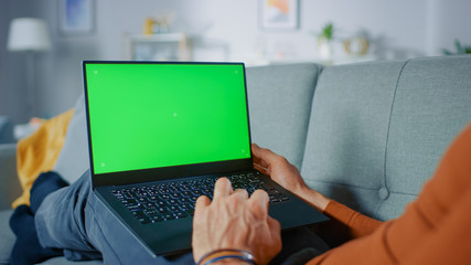 Man Lying on His Sofa At Home, Uses Laptop with Green Mock-up Screen. Over The Shoulder Shot of Anonymous Man in the Living Room.