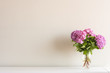 © Natalie - Pink hydrangea flowers with green leaves in glass vase on white side board against neutral wall background