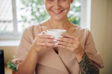 Cropped image of smiling young woman drinking cup of delicious black coffee after finishing work