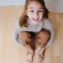 Wall Mural - Little girl in a beige knitted sweater sitting on a parquet at home. She's holding her bare feet with hands. Making grimace with wide open eyes and mouth. Top view.