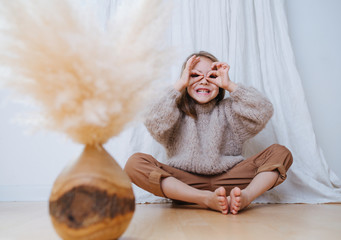 Wall Mural - Smiling little girl in a beige knitted sweater sitting in a butterfly yoga position on a parquet at home, in front of a curtain. She making grimace with grin and hand goggles.