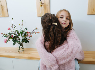 Wall Mural - Two little girls in pink knitted sweaters hugging in a corridor. One is sitting on the shelf and looking at camera. Next to cut roses in a jar.