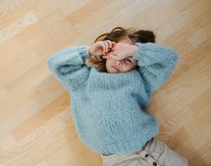 Wall Mural - Sleepy little girl in a blue knitted sweater waking up from napping on the floor. She cover's her eye from the bright light. At home, top view.