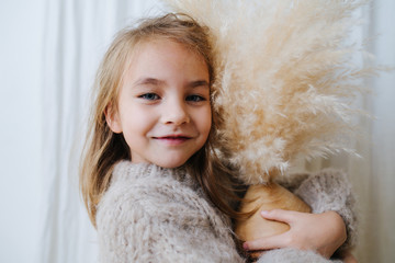 Wall Mural - Portrait of a little girl in a beige knitted sweater hugging coconut vase with cereal bouquet in it. Soft fluffy spikes touching her cheek. At home, in front of a curtain.
