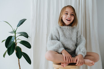 Wall Mural - Happy little barefoot girl in a grey knitted sweater sitting cross-legged on a stool at home, in front of a curtain next to a potted plant.