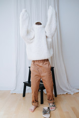 Wall Mural - Funny little girl struggling to put on white fluffy knitted sweater. It does not slide down on inself, so her head and arms still inside. At home, in front of a curtain. Full length.