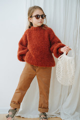 Wall Mural - Little girl in dark orange knitted sweater, round sunglasses, brown jeans, leopard sandals with net bag. She's posing for photo, trying to look cute and fashionable. One leg on toes. Full length.