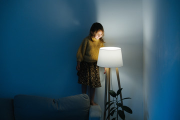 Wall Mural - Brunette little girl in yellow knitted sweater and leopard skirt standing on a backseat of a couch at home. She's looking at dim night light from above. Her face is lit.