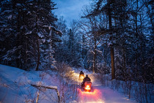 Snowmobile In The Evening Goes Through The Winter Forest. Headlights. Night Road Through The Winter Forest. Snowmobile At Night