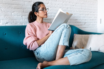 Pretty young woman reading a book while sitting on sofa at home.