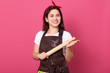 © sementsova321 - Happy chef woman holding rolling pin in hands isolated over pink background, smiling housewife wearing brown apron, white shirt and red head band looking at camera, female being in good mood.